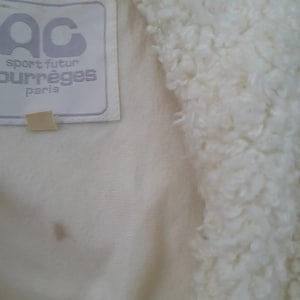 May include: Close-up of a cream-colored jacket with a fluffy, textured collar. The jacket features a label with the text "AC sport futur courr&egrave;ges paris". A small, tan tag is attached below the label. The fabric has a soft, plush appearance.