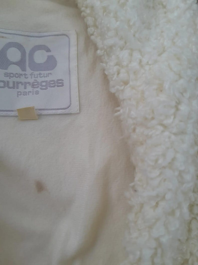 May include: Close-up of a cream-colored jacket with a fluffy, textured collar. The jacket features a label with the text "AC sport futur courr&egrave;ges paris". A small, tan tag is attached below the label. The fabric has a soft, plush appearance.