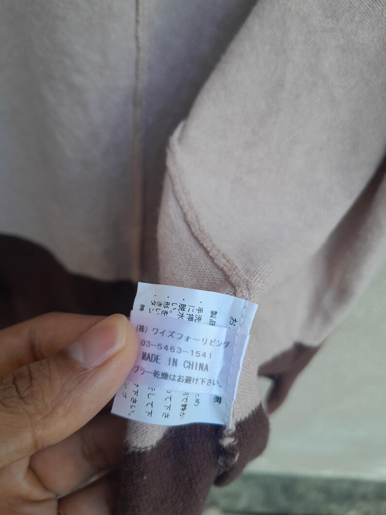 K&ouml;nnte beinhalten: Beige-braunes Kleidungsst&uuml;ck mit einem Etikett mit der Aufschrift "MADE IN CHINA". Das Etikett enth&auml;lt auch japanischen Text und eine Telefonnummer. Das Kleidungsst&uuml;ck hat ein zweifarbiges Design.