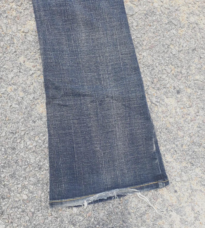 Puede incluir: Primer plano de una secci&oacute;n de vaqueros de mezclilla azul oscuro. Los vaqueros tienen un aspecto ligeramente descolorido y un dobladillo deshilachado. La tela muestra patrones de tejido verticales y horizontales.