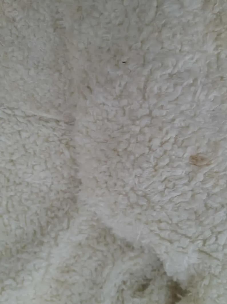 May include: Close-up of a cream-colored, textured fabric with a dense, fluffy appearance. The material has a soft, plush texture, with slight variations in color and small dark specks.