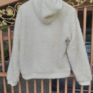 May include: Cream-colored, fluffy hoodie with a hood and long sleeves. The soft, textured material is visible, and the cuffs and hem are ribbed. The hoodie is hanging on a wooden hanger.