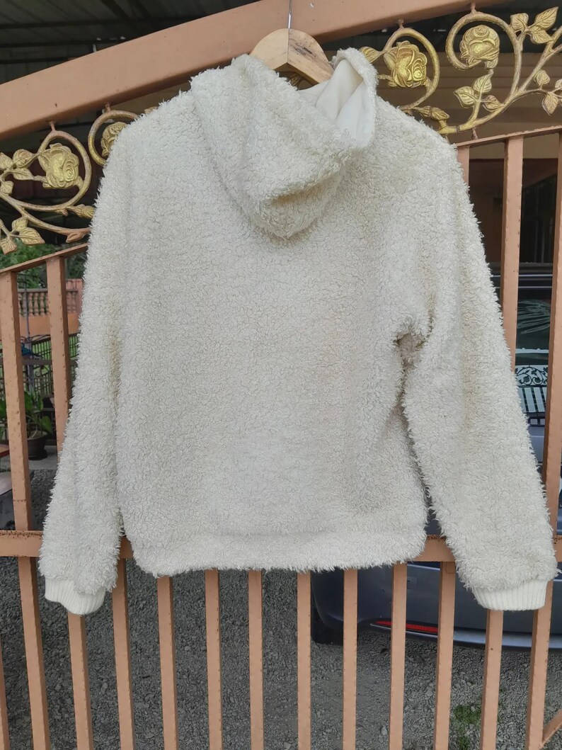 May include: Cream-colored, fluffy hoodie with a hood and long sleeves. The soft, textured material is visible, and the cuffs and hem are ribbed. The hoodie is hanging on a wooden hanger.