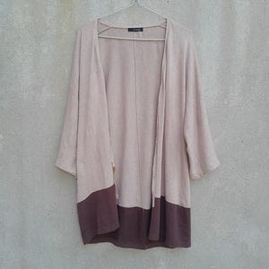 K&ouml;nnte beinhalten: Ein hellbrauner offener Cardigan mit Dreiviertel&auml;rmeln. Der untere Teil des Cardigans ist dunkelbraun. Der Cardigan h&auml;ngt an einem Metallb&uuml;gel vor einer hellgrauen Wand.