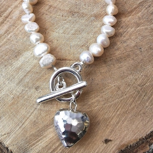 Peut inclure: Un bracelet en perles blanches avec un fermoir à glissière argenté et un pendentif en forme de cœur en argent martelé.