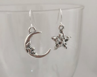 May include: A pair of silver earrings with a crescent moon and a star charm. The earrings are hanging from a glass surface.