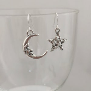 May include: A pair of silver earrings with a crescent moon and a star charm. The earrings are hanging from a glass surface.