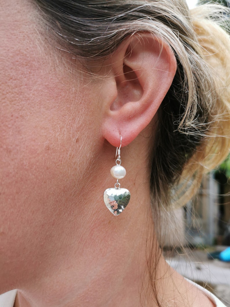 May include: Silver heart earrings with a white pearl dangle. The heart is hammered and has a textured surface.
