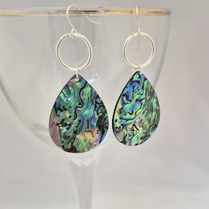 Op de afbeelding: Oorbellen met iriserende abalone schelpen hangers. De druppelvormige hangers tonen wervelende patronen in groen, blauw en paars. Ze zijn bevestigd aan zilverkleurige ringen en oorhaken, wat een opvallend contrast biedt.