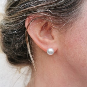 May include: A close-up shot of a person's ear with a large, round, light gray pearl stud earring. The person's hair is pulled back, revealing the ear and earring. The earring is a simple, elegant design.