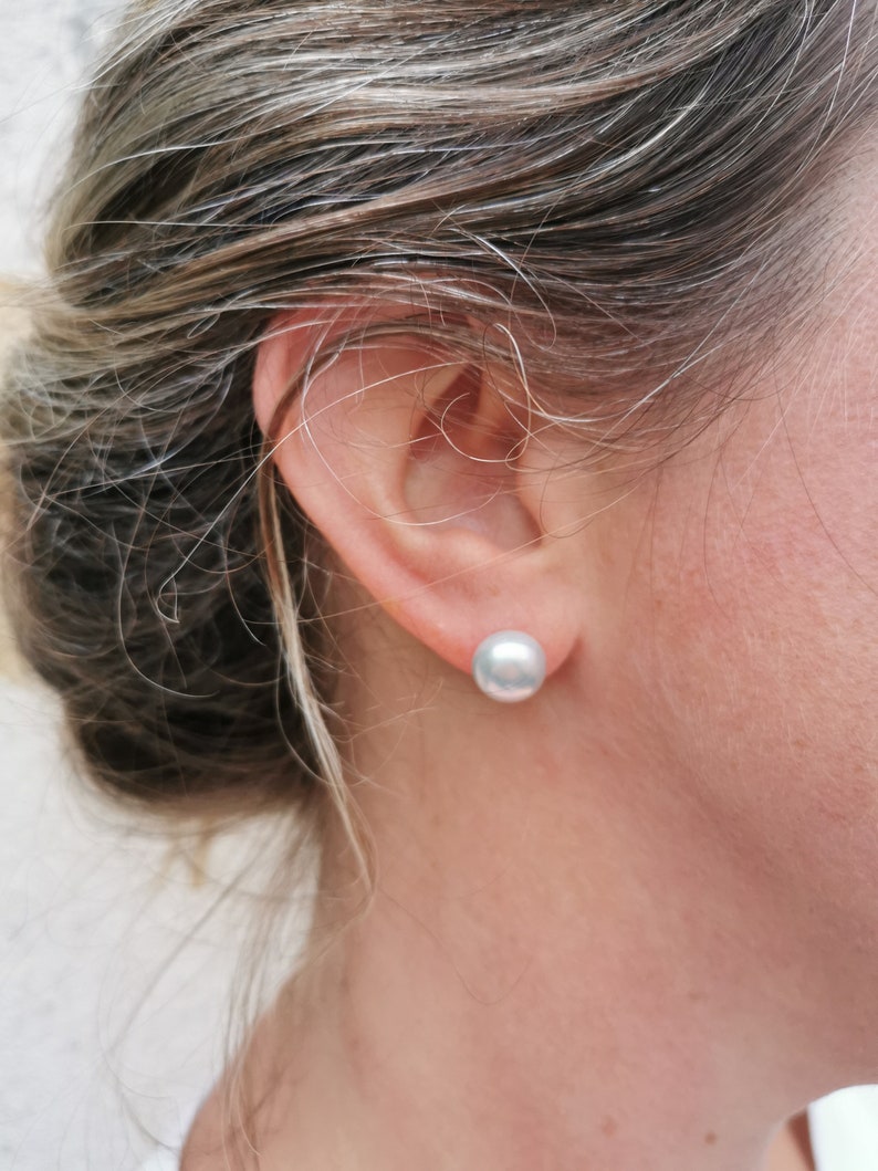 May include: A close-up shot of a person's ear with a large, round, light gray pearl stud earring. The person's hair is pulled back, revealing the ear and earring. The earring is a simple, elegant design.