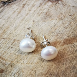 May include: A pair of white pearl stud earrings with silver posts. The pearls are round and have a smooth, lustrous surface. The earrings are displayed on a light brown, textured wooden surface.
