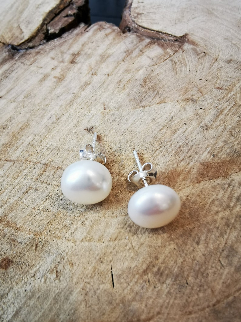 May include: A pair of white pearl stud earrings with silver posts. The pearls are round and have a smooth, lustrous surface. The earrings are displayed on a light brown, textured wooden surface.
