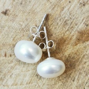 May include: A pair of oval-shaped, white pearl stud earrings with silver posts and backs. The pearls have a smooth, lustrous surface, reflecting light. The earrings are displayed on a light brown, textured surface.