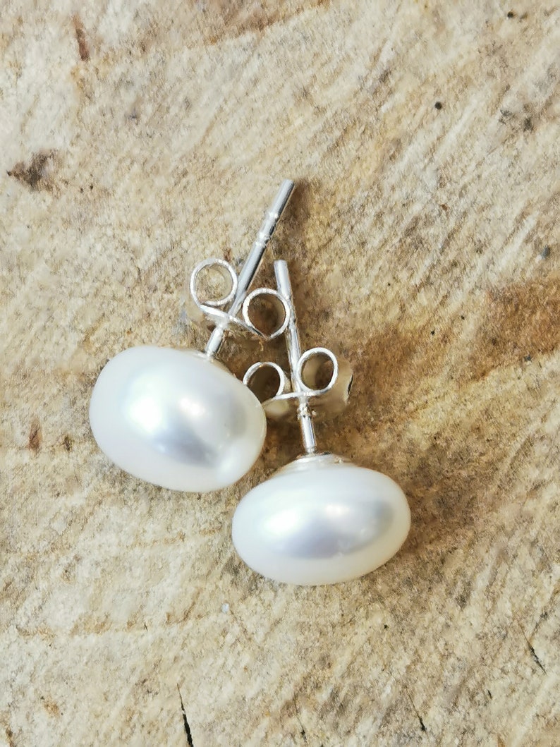 May include: A pair of oval-shaped, white pearl stud earrings with silver posts and backs. The pearls have a smooth, lustrous surface, reflecting light. The earrings are displayed on a light brown, textured surface.