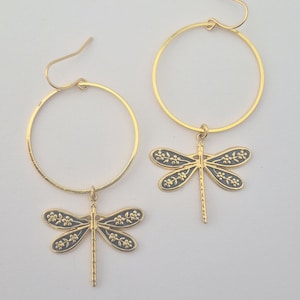 Può includere: Un paio di orecchini a cerchio color oro con ciondoli a forma di libellula. I ciondoli a forma di libellula sono grigio scuro con dettagli floreali color oro sulle ali. Sfondo bianco.