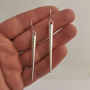 May include: A pair of silver earrings with long, pointed, and tapered designs. The earrings are hanging from a person's hand.