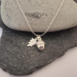 May include: Silver necklace with an acorn pendant and oak leaf charm. The acorn pendant is approximately 2 cm high. The chain is fine, and the necklace is displayed on a dark grey, textured stone surface, creating a natural aesthetic.