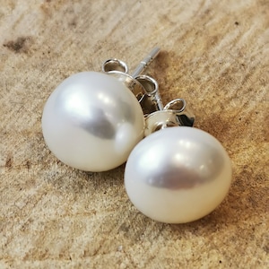 May include: A pair of white pearl stud earrings with silver posts and backings. The pearls are round and have a lustrous sheen. The earrings are set against a light brown, textured background.