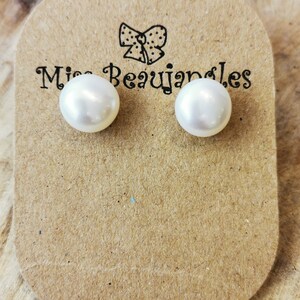 May include: A pair of white pearl stud earrings on a brown card with the text "Miss Beaujangles" and a bow illustration. The earrings are round and have a glossy finish. The card is placed on a wooden surface.