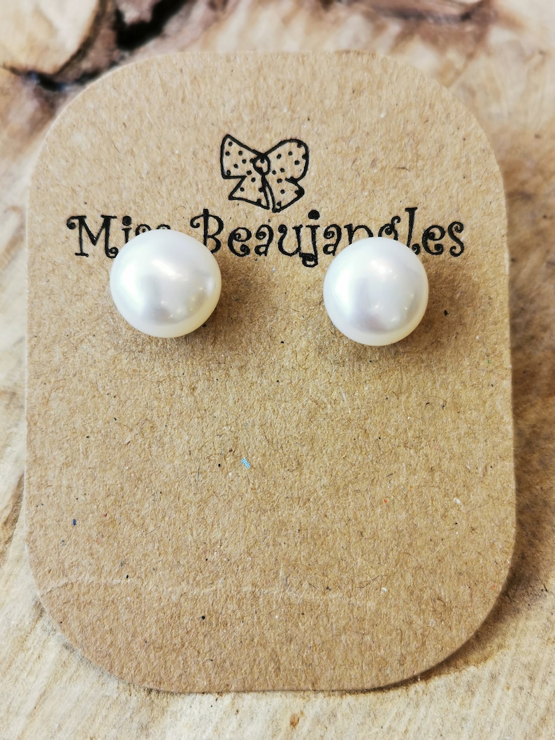 May include: A pair of white pearl stud earrings on a brown card with the text "Miss Beaujangles" and a bow illustration. The earrings are round and have a glossy finish. The card is placed on a wooden surface.