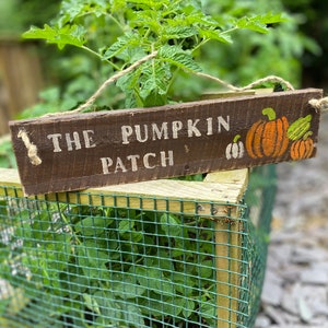 May include: A wooden sign with the text "THE PUMPKIN PATCH" painted in white. The sign is hanging from a tomato plant in a garden. The sign has a painted image of pumpkins.