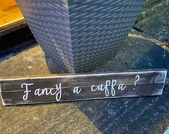 May include: A rustic wooden sign with white painted lettering that reads "Fancy a cuppa?"