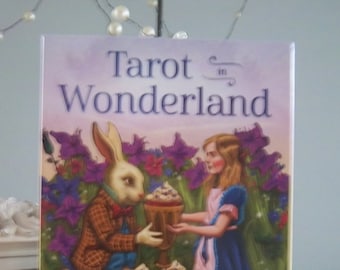 May include: Tarot in Wonderland deck box featuring a white rabbit in a waistcoat holding a cup with a girl in a blue dress. The box has a purple, pink, and green background with flowers and the text "Tarot in Wonderland" and "Barbara Moore Illustrated by Eugene Smith".