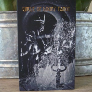 May include: A tarot card deck titled "Circle of Doors Tarot" with a black and white illustration. The image features a mystical figure with antlers, a key, and a swirling, ethereal background. The text is in gold.