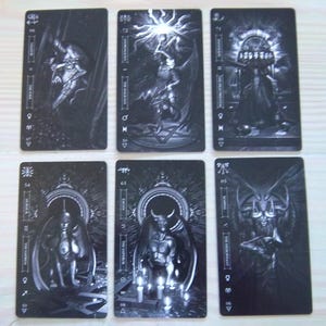 May include: Six black and white tarot cards are displayed, each featuring intricate illustrations of figures and symbols. The cards have a rectangular shape with rounded corners. The images include figures with wings, demons, and other mystical imagery. The cards are arranged on a light-colored surface.