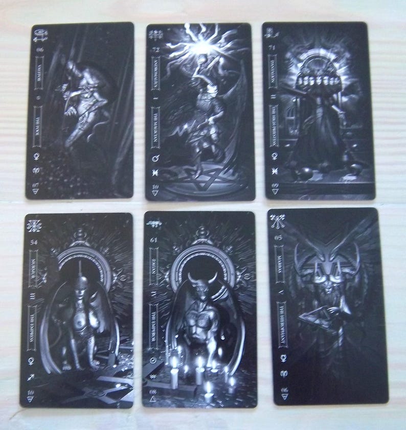 May include: Six black and white tarot cards are displayed, each featuring intricate illustrations of figures and symbols. The cards have a rectangular shape with rounded corners. The images include figures with wings, demons, and other mystical imagery. The cards are arranged on a light-colored surface.