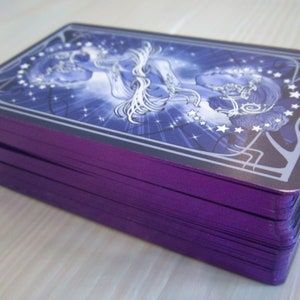 May include: A deck of tarot cards with a purple back and a blue and white illustration of a woman with stars on the front.