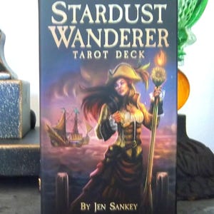 以下が含まれることがあります： 「Stardust Wanderer Tarot Deck」の箱は、黒い背景に金色の文字が特徴です。イラストには、海賊帽をかぶり、炎が灯った杖を持つ人物が描かれており、背景には木製の桟橋と帆船が見えます。