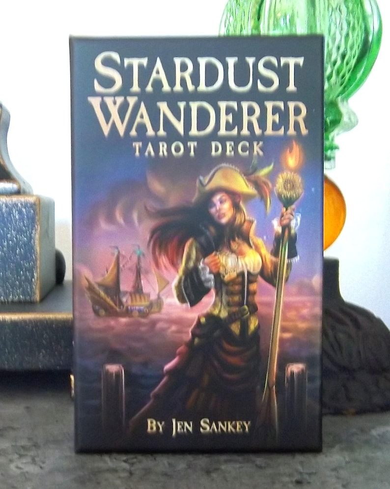 以下が含まれることがあります： 「Stardust Wanderer Tarot Deck」の箱は、黒い背景に金色の文字が特徴です。イラストには、海賊帽をかぶり、炎が灯った杖を持つ人物が描かれており、背景には木製の桟橋と帆船が見えます。