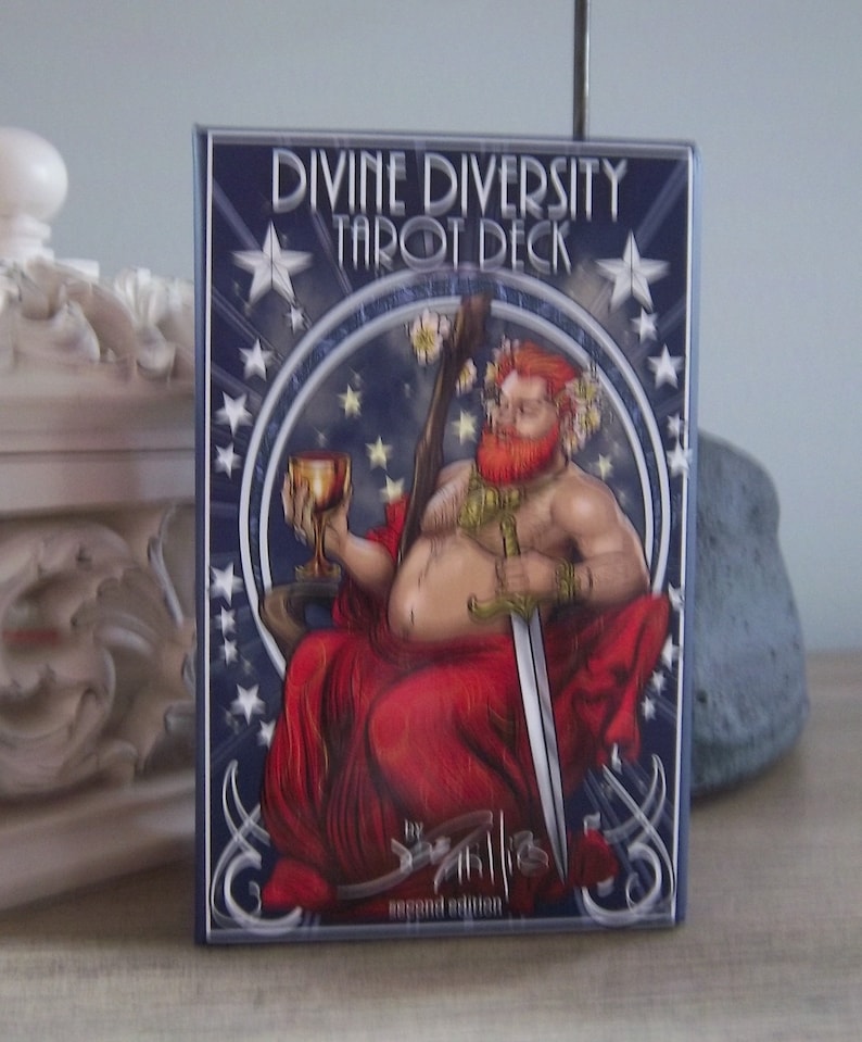May include: A blue and silver tarot deck box with the title "Divine Diversity Tarot Deck" and a depiction of a red-haired, bearded figure holding a sword and a chalice. The box also includes the text "Second Edition".
