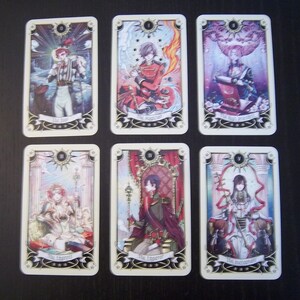 May include: A set of ten tarot cards featuring illustrations of people in various poses. The cards are numbered from 0 to 9 and have gold trim. The cards are on a light brown surface.