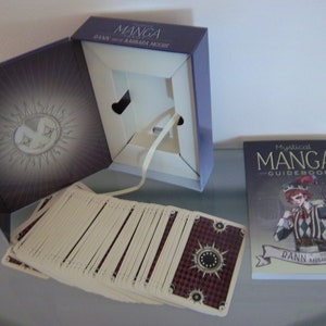 May include: A Mystical Manga Guidebook with a deck of cards in a purple box. The guidebook features a black and white illustration of a jester with red hair and a black and white checkered outfit. The box has a sun with a face on it.