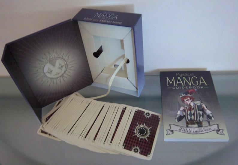 May include: A Mystical Manga Guidebook with a deck of cards in a purple box. The guidebook features a black and white illustration of a jester with red hair and a black and white checkered outfit. The box has a sun with a face on it.