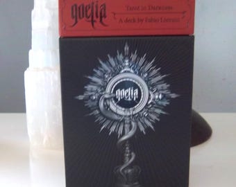 Puede incluir: Una caja negra de cartas del tarot con la palabra "goetia" en una fuente gótica y una caja roja encima. La caja presenta una detallada ilustración plateada de una serpiente y un diseño circular. El texto "Tarot in Darkness" es visible.