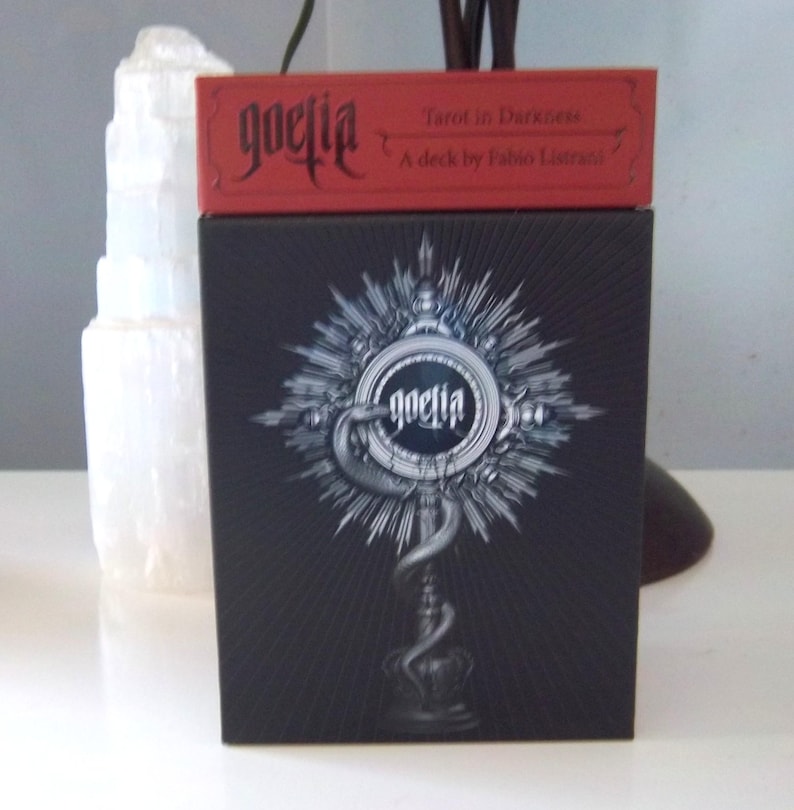 May include: A black tarot card box with the word "goetia" in a gothic font, and a red box on top. The box features a detailed silver illustration of a serpent and a circular design. The text "Tarot in Darkness" is visible.