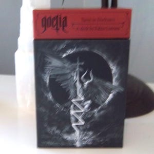 May include: A black box with a white illustration of a winged figure with horns and a cross. The box has a red lid with the word "goetia" and the text "Twist in Darkness".