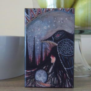 May include: A rectangular art print depicting a mystical scene. A woman with long black hair holds a moon-like orb. A large black bird with red eyes and intricate patterns is set against a starry night sky. The colour palette includes blues, blacks, and reds.