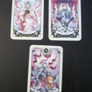 May include: Three tarot cards with illustrations of angels, a knight, and a woman with a lion. The cards are white with gold trim and feature the Roman numeral and name of the card. The cards are on a dark surface.