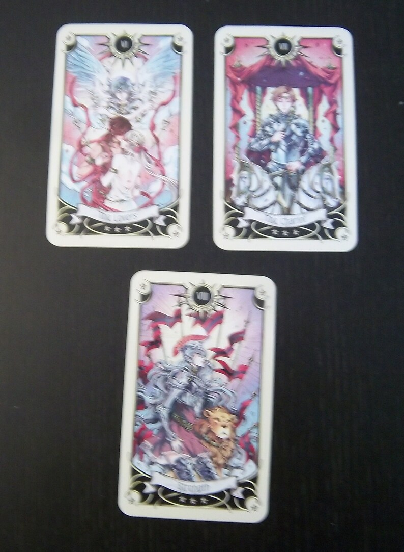 May include: Three tarot cards with illustrations of angels, a knight, and a woman with a lion. The cards are white with gold trim and feature the Roman numeral and name of the card. The cards are on a dark surface.