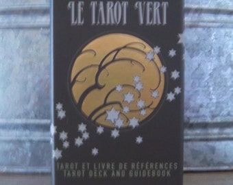Może przedstawiać: Szare pudełko z napisem "LE TAROT VERT" w białym kolorze. Pudełko zawiera złote koło ze stylizowanym drzewem i białymi gwiazdami. Tekst "TAROT ET LIVRE DE REFERENCES TAROT DECK AND GUIDEBOOK" jest nadrukowany na biało pod kołem. Tekst "L'HERMITE" jest nadrukowany na biało na dole pudełka.