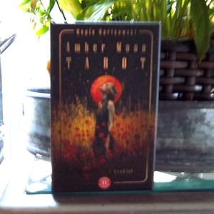 Può includere: Una scatola di mazzo di carte dei tarocchi con il titolo "Amber Moon Tarot" in lettere dorate. La scatola presenta un'illustrazione di una donna in piedi in un campo di fiori rossi con un grande sole arancione sullo sfondo. La scatola è scura con un bordo dorato.