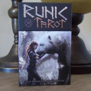 Op de afbeelding: Een tarotkaartspel getiteld "RUNIK TAROT" met een zwarte bovenkant en een fantasie-illustratie. De illustratie toont een vrouw met rood haar en een ijsbeer in een bosrijke omgeving. De auteurs van het deck zijn Jack Sephiroth en Allen Dempster.