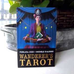 May include: A box for Wanderer's Tarot cards, featuring a colorful illustration of a person in a purple jacket and a hat, sitting in a meditative pose. The box is black with gold lettering that reads "Wanderer's Tarot" and "Pierluca Zizzi + Michele D'Aloisio".