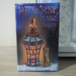 May include: A box of Reverse Scale Tarot cards with a blue and gold design. The box features a lantern with a lit candle inside and a stick figure standing next to it. The text on the box reads "Reverse Scale Tarot" and "78 Tarot Cards & Guidebook".