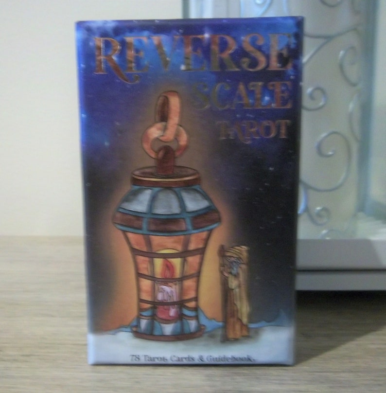 May include: A box of Reverse Scale Tarot cards with a blue and gold design. The box features a lantern with a lit candle inside and a stick figure standing next to it. The text on the box reads "Reverse Scale Tarot" and "78 Tarot Cards & Guidebook".